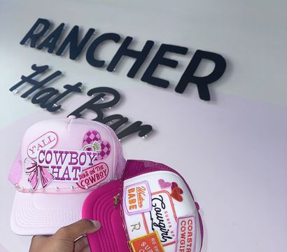 Hand holding two pink embroidered trucker hats with cowboy and cowgirl patches, hearts and chain accents, photographed inside a hat shop under a black wall sign.