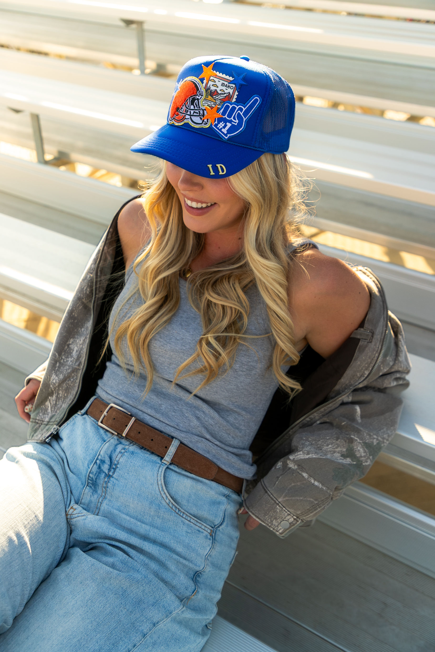 Smiling woman wearing a blue Idaho baseball cap, gray tank top, camo jacket and high-waist jeans sitting on sunlit metal bleachers.