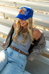 Smiling woman wearing a blue Idaho baseball cap, gray tank top, camo jacket and high-waist jeans sitting on sunlit metal bleachers.