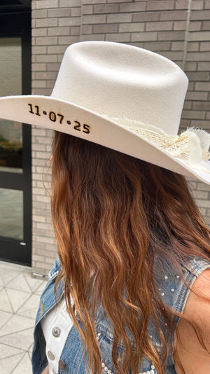Person with long wavy brown hair wearing a white cowboy hat stamped “11·07·25” and a studded denim vest standing by a brick building entrance