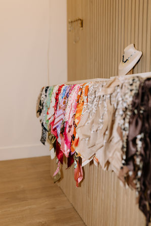 Vibrant patterned silk scarves cascading over a corrugated wooden counter in a boutique display, with a necklace-styled bust on top and warm hardwood floor beneath.