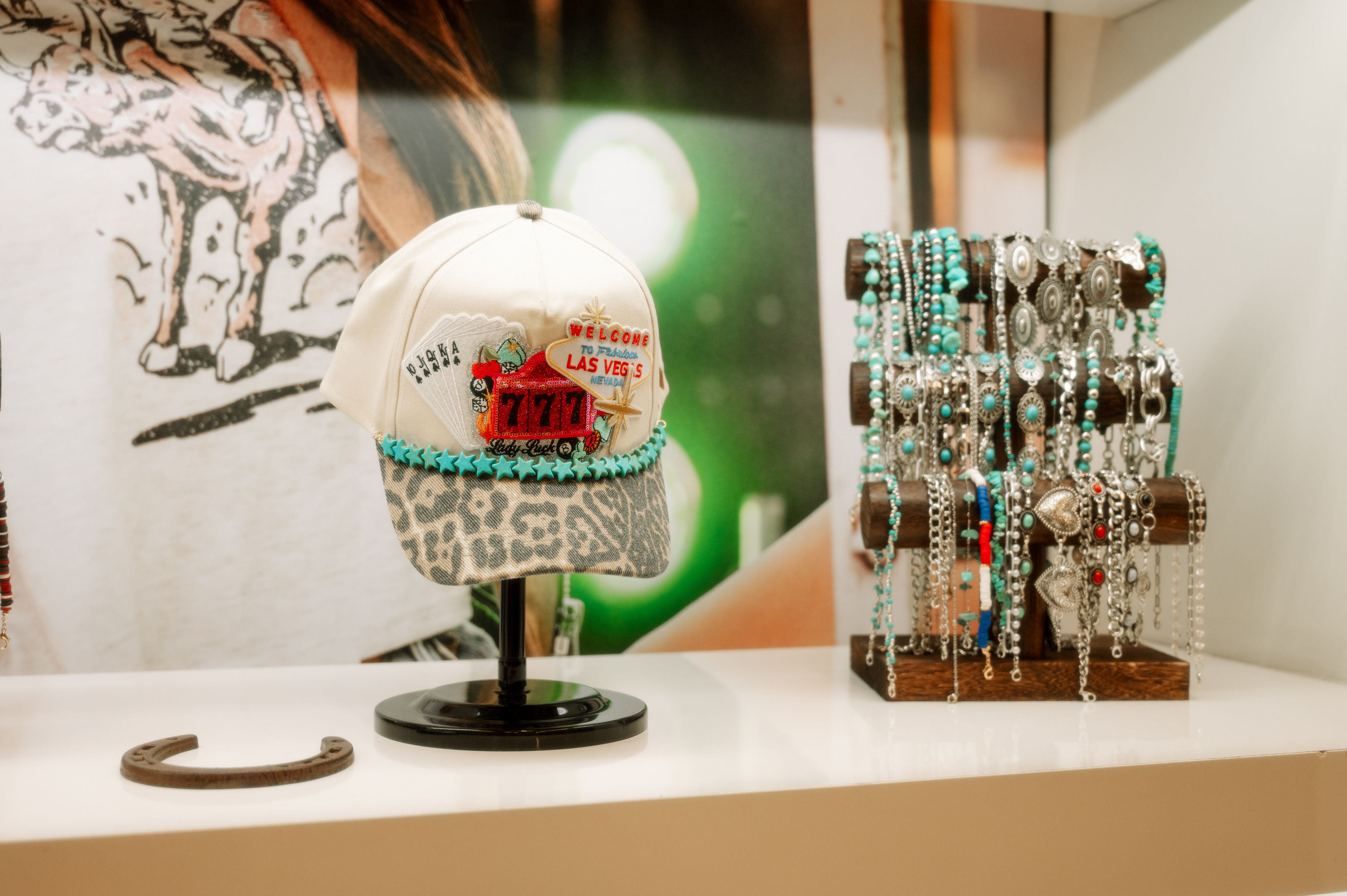 White trucker hat with leopard-print brim, turquoise star trim and a Las Vegas 777 patch on a display stand beside a wooden rack of turquoise and silver bracelets on a retail shelf.