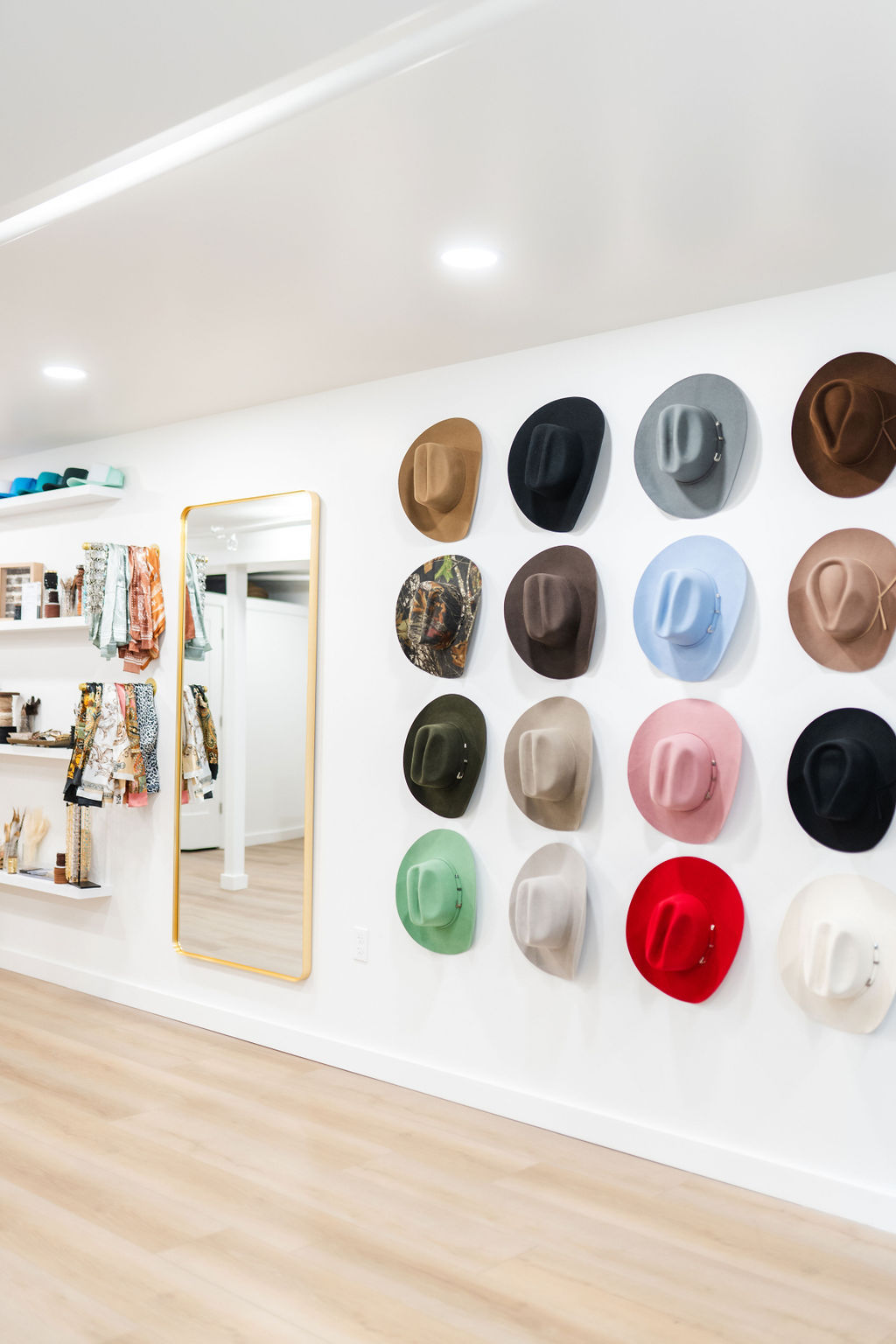 Bright modern boutique interior with a wall display of colorful felt hats arranged in neat rows beside a gold-framed full-length mirror and shelves of scarves and accessories on light wood floors.