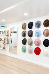Bright modern boutique interior with a wall display of colorful felt hats arranged in neat rows beside a gold-framed full-length mirror and shelves of scarves and accessories on light wood floors.