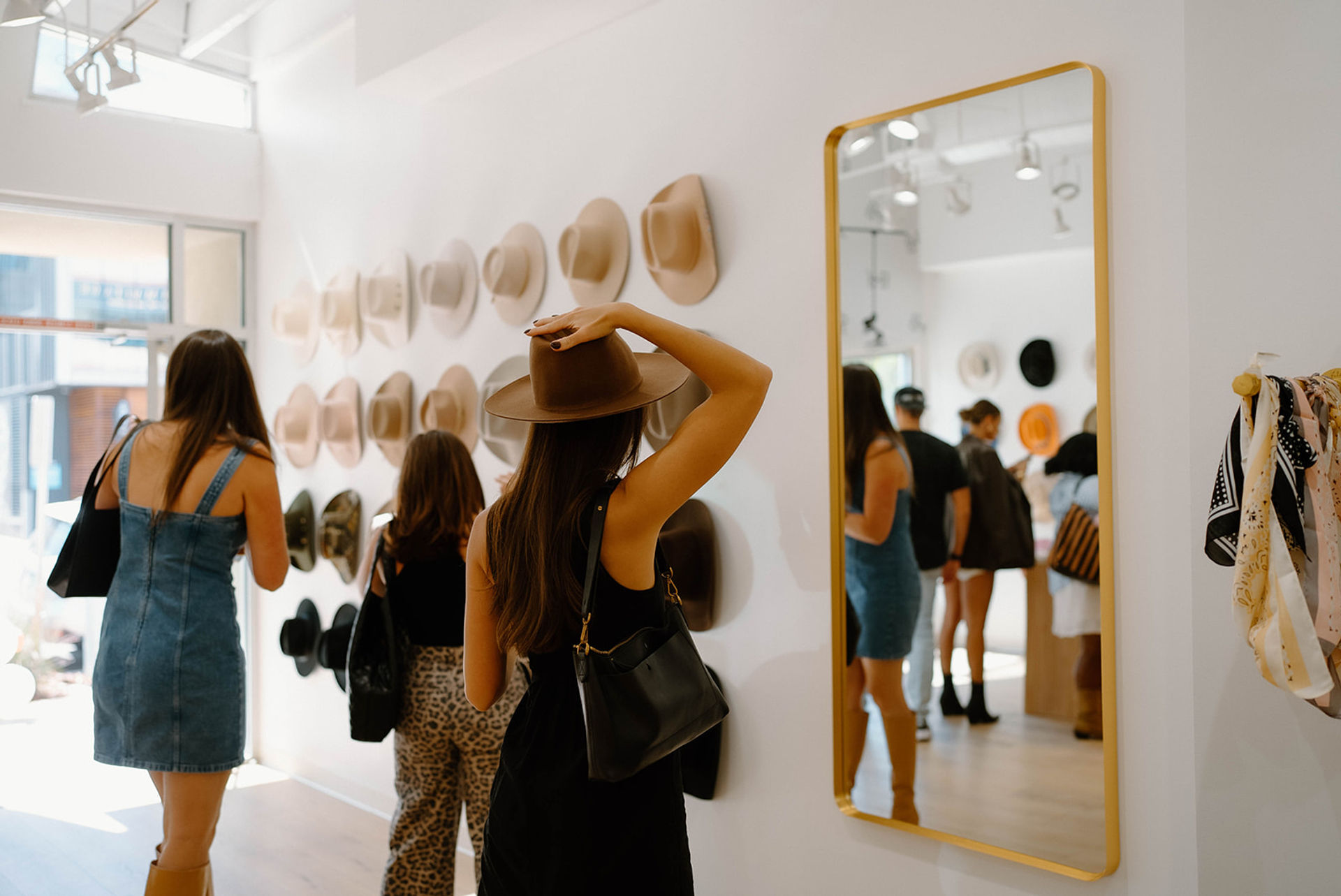 Sunlit boutique interior with women browsing wall-mounted wide-brim hats; one woman tries on a brown hat in front of a gold-framed mirror while others explore hats and scarves.