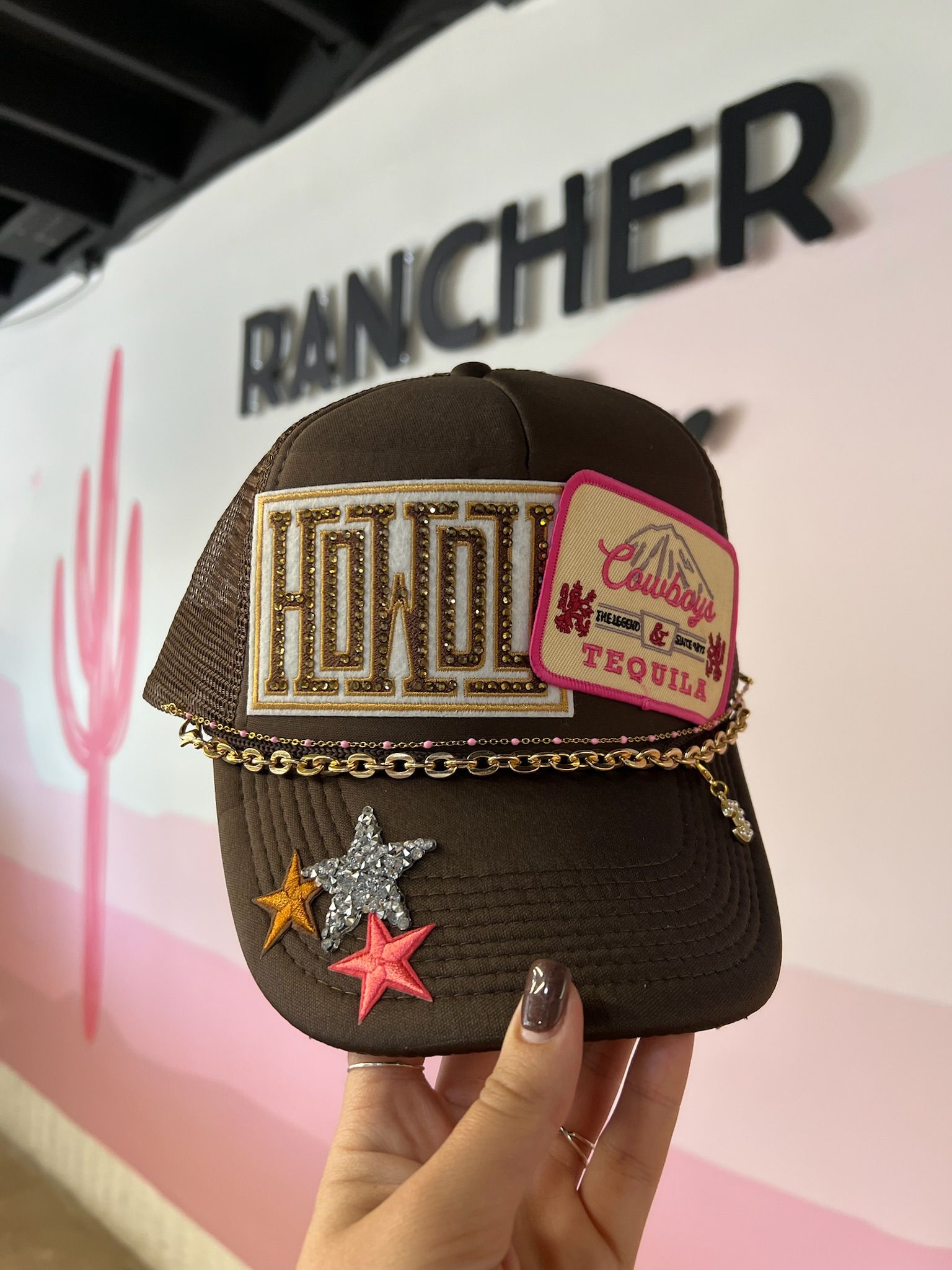 Brown embellished trucker hat held by hand with gold-studded embroidered patch, pink “Tequila” patch, gold chain trim and glitter star appliqués against a pink-and-white boutique wall.
