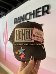 Brown embellished trucker hat held by hand with gold-studded embroidered patch, pink “Tequila” patch, gold chain trim and glitter star appliqués against a pink-and-white boutique wall.