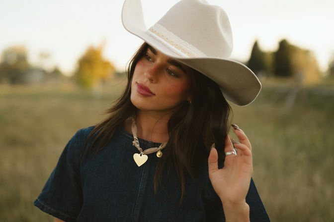 Woman in beige cowboy hat and denim top standing in a rural field at golden hour, touching her hair and wearing a gold heart pendant — relaxed western-style portrait.