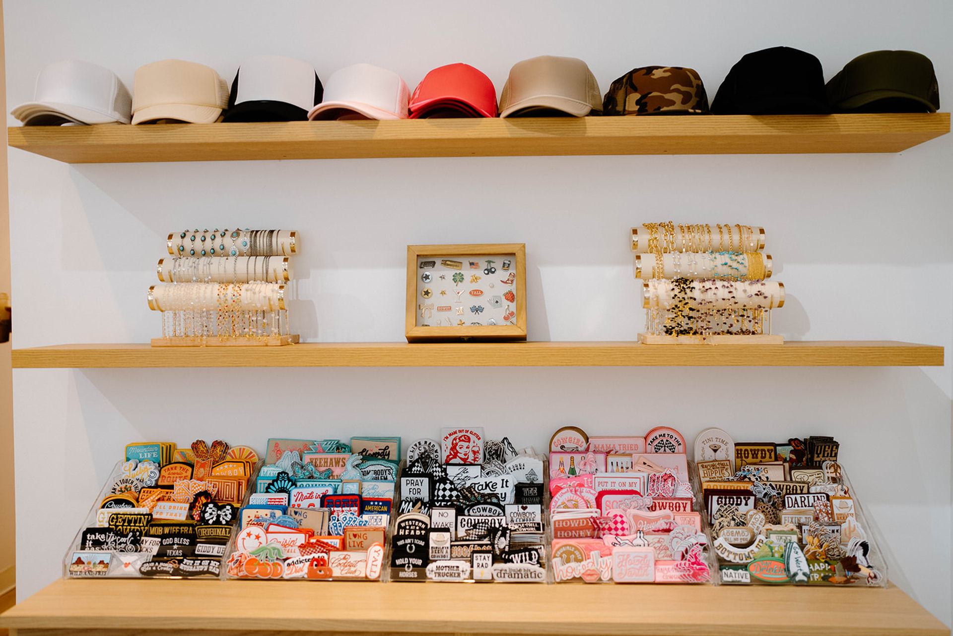 Wooden wall shelves in a boutique displaying a row of colorful baseball caps on the top shelf, bracelets and a framed pin set on the middle shelf, and organized trays of vibrant enamel pins, patches and stickers on the bottom shelf.