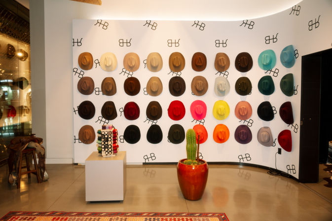 Bright boutique interior with a playful rainbow of felt cowboy hats mounted in a grid on a white wall, terracotta pot cactus in front and a small pedestal display of accessories on the polished floor.