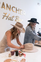 Two women in cowboy-style hats customize beige and black wide-brim felt hats at a hat-styling counter, surrounded by ribbons, trims, scissors and other hat-making tools in a western hat workshop scene.