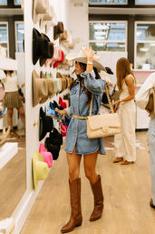 Woman in a denim dress tries on a white cowboy hat in a boutique hat shop, standing by a wall of colorful hats while wearing brown cowboy boots and a beige quilted shoulder bag.