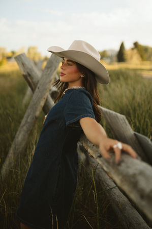 Young woman in a beige cowboy hat and denim dress leaning on a rustic wooden fence in a grassy country field at golden hour, relaxed cowgirl vibe