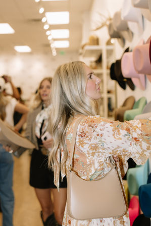 Blonde woman in a floral dress with a beige shoulder bag browsing colorful hats on a wall display inside a bright boutique