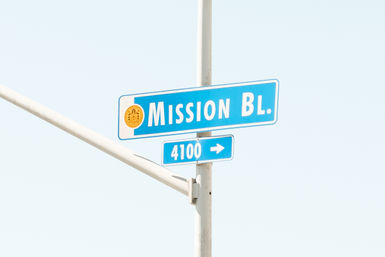 Sunlit blue 'Mission Bl.' street sign with a '4100' block and arrow mounted on a metal pole against a clear pale sky
