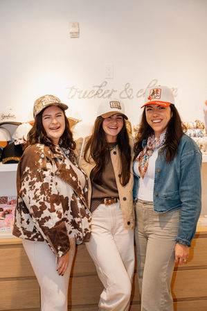 Three smiling women in trucker hats and western-inspired outfits pose together inside a boutique hat display.