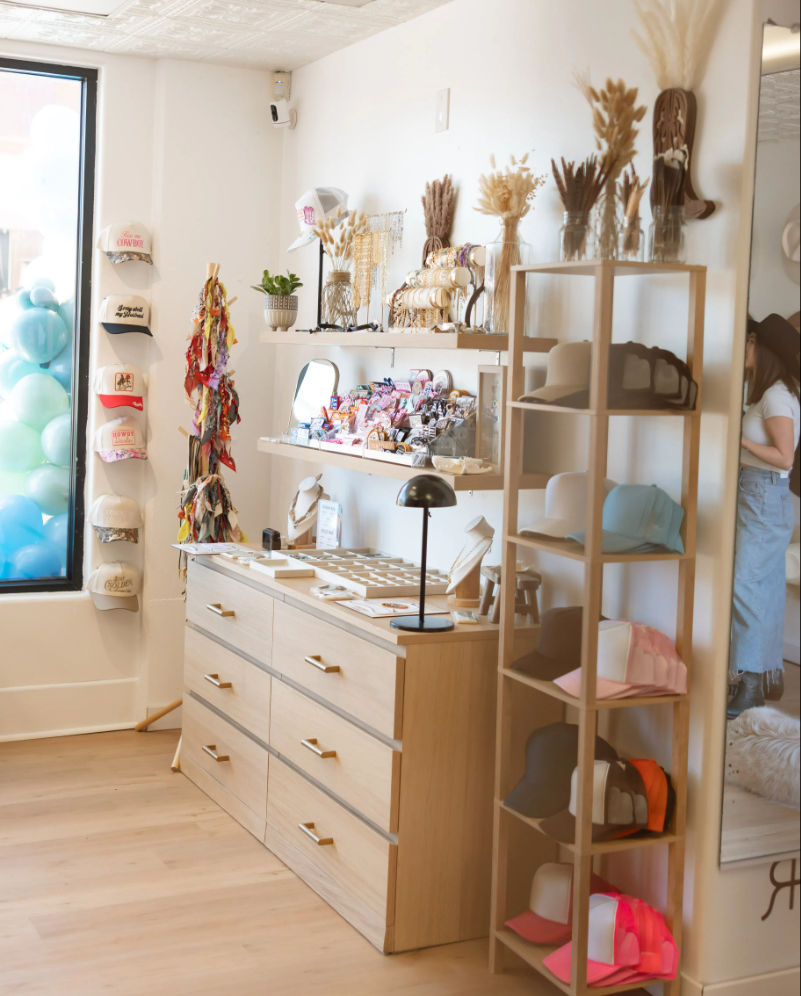 Bright boutique interior with light wood drawers and open shelving displaying caps, jewelry, colorful accessories and dried floral arrangements beside a storefront window with pastel balloons