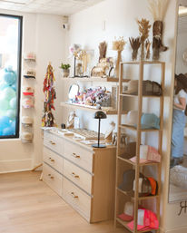 Bright boutique interior with light wood drawers and open shelving displaying caps, jewelry, colorful accessories and dried floral arrangements beside a storefront window with pastel balloons