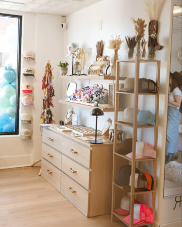 Bright boutique interior with light wood drawers and open shelving displaying caps, jewelry, colorful accessories and dried floral arrangements beside a storefront window with pastel balloons