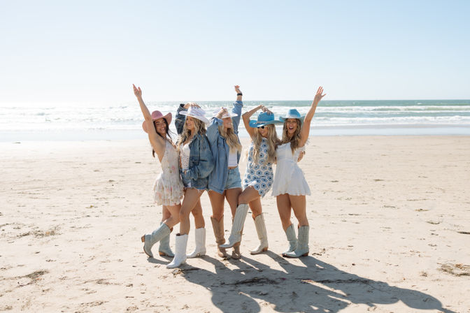 Five friends in summer dresses, cowboy boots and colorful wide-brim hats cheer and pose together on a sunny sandy beach with ocean waves in the background.