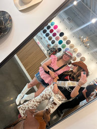 Mirror selfie in a western hat boutique — two people trying on cowboy hats in front of a colorful hat wall, white cowboy boots, cow-print pants and a leather saddle in foreground.