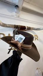Brown felt western cowboy hat with braided leather band, feathers, ribbon and vintage playing cards tucked in, held up against a white wall with decorative longhorn horns in a boutique interior.