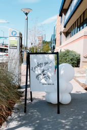 A-frame sidewalk sign reading 'Cowboy up!' with a bucking-horse illustration and a cluster of white balloons beside it on a sunny sidewalk near a parking meter and storefront in Cherry Creek, CO.