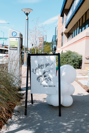 A-frame sidewalk sign reading 'Cowboy up!' with a bucking-horse illustration and a cluster of white balloons beside it on a sunny sidewalk near a parking meter and storefront in Cherry Creek, CO.