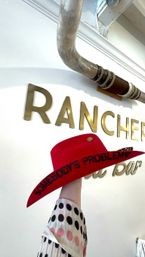 Hand lifts a bright red cowboy hat with "SOMEBODY'S PROBLEM" stitched on the brim, in a western‑themed shop interior with gold wall letters and a rustic overhead pipe.