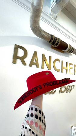 Hand lifts a bright red cowboy hat with "SOMEBODY'S PROBLEM" stitched on the brim, in a western‑themed shop interior with gold wall letters and a rustic overhead pipe.