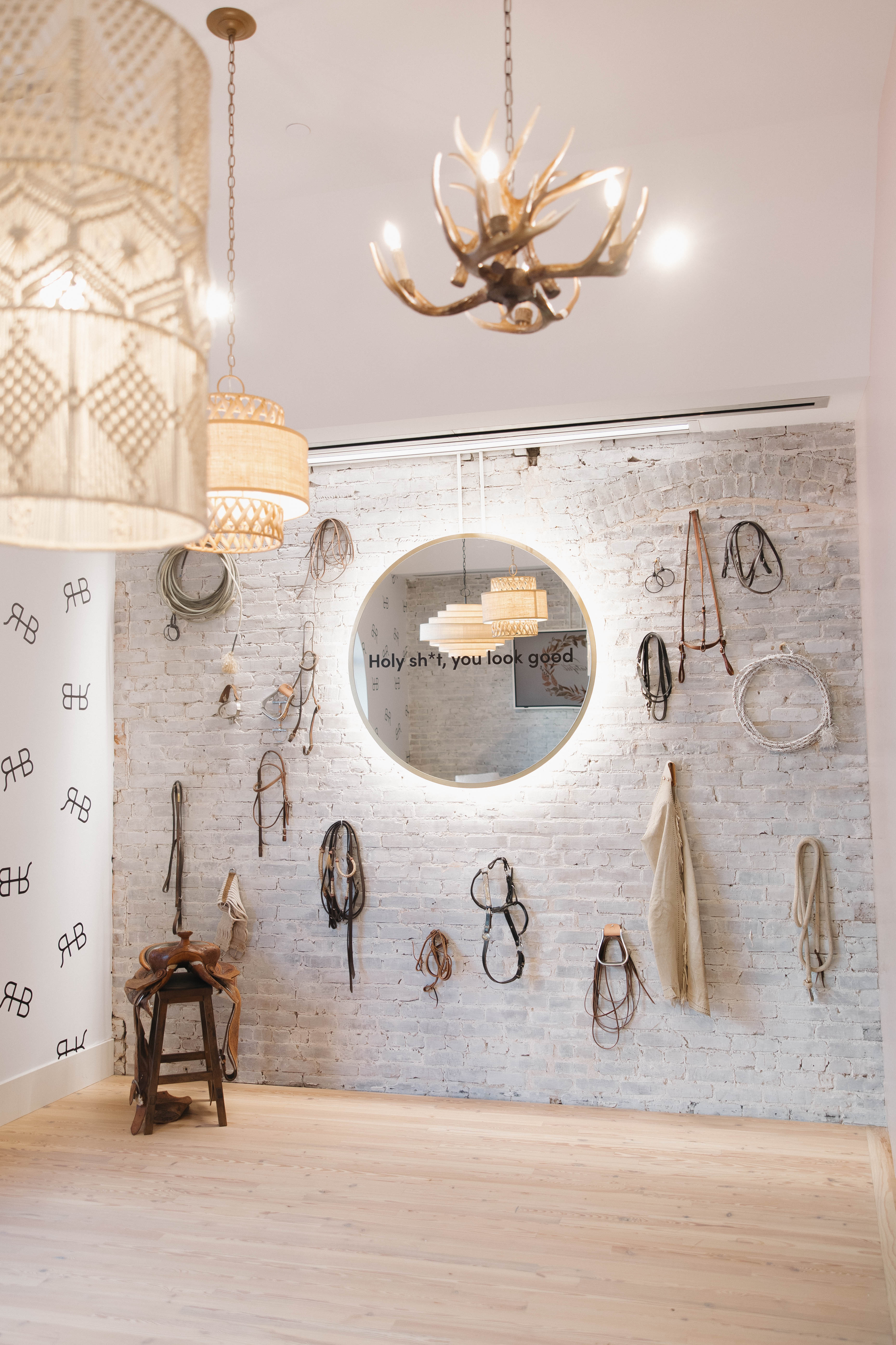 Bright boutique-style interior with whitewashed brick wall, round backlit mirror reading 'Holy sh*t, you look good,' hanging horse tack and ropes, a saddle on a stool, woven pendant lamps and antler chandelier above pale wood floor