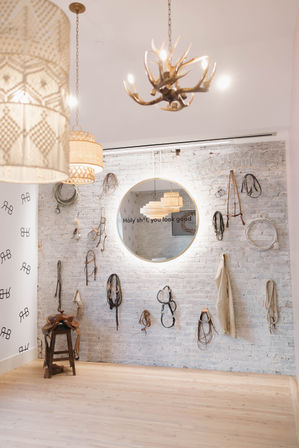 Bright boutique-style interior with whitewashed brick wall, round backlit mirror reading 'Holy sh*t, you look good,' hanging horse tack and ropes, a saddle on a stool, woven pendant lamps and antler chandelier above pale wood floor