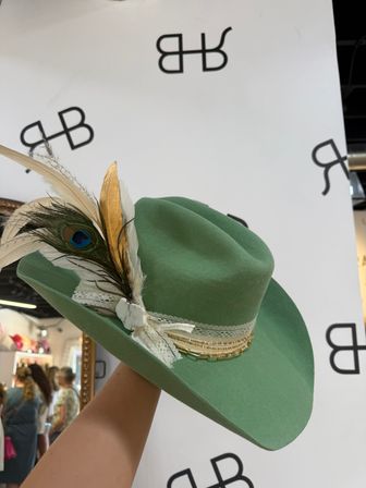 Handheld mint green felt cowboy hat with wide brim, decorated with peacock and gold feathers, lace ribbon, marble-look pin and layered gold chain trim, photographed in a boutique