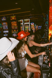 Three friends in cowboy hats and western outfits laughing and playing glowing slot machines inside a lively casino nightlife scene.