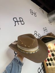 Brown felt cowboy hat with layered ribbon band, turquoise bead trim and dangling gold star chain, held by a hand in a denim sleeve against a white logo‑patterned boutique wall.