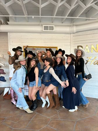 Smiling group of women posing playfully in cowboy hats and western boots inside a bright rustic hat boutique with white shiplap walls and hat displays.
