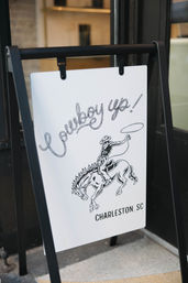 A black A-frame sidewalk sign outside a storefront in Charleston, SC featuring a white poster with a hand-drawn cowboy on a bucking horse and rope-style lettering.