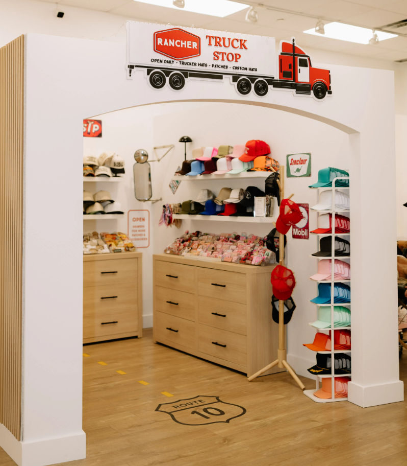 Playful truck-stop themed retail nook seen through an archway, filled with colorful baseball caps on shelves and a rotating rack, light wood drawer cabinets, Route 10 road decal on the hardwood floor, and retro gas-station style signs.