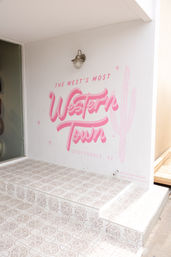 Cheery pink script mural reading “Western Town” with a cactus illustration on a white stucco wall above patterned tile steps at an entryway in Scottsdale, Arizona.