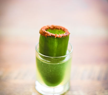 Zesty chili-rimmed cucumber shot — vibrant green fresh cucumber juice served in a small glass with a hollow cucumber garnish, on a wooden table