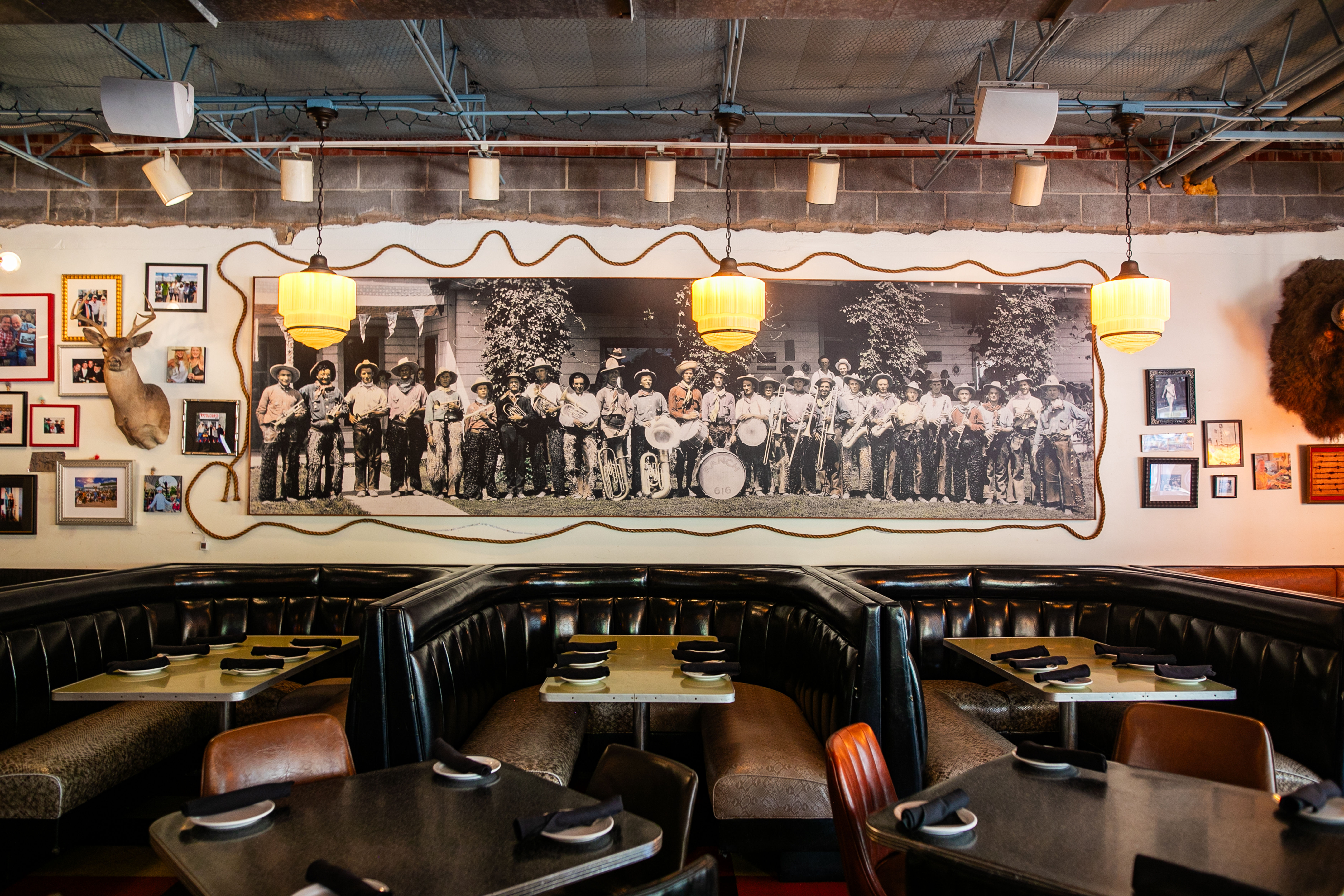 Vintage Western-style diner interior with curved black leather booths and small tables set for service, large sepia group photo mural of a band on the wall, hanging pendant lights, framed photos and a mounted deer head