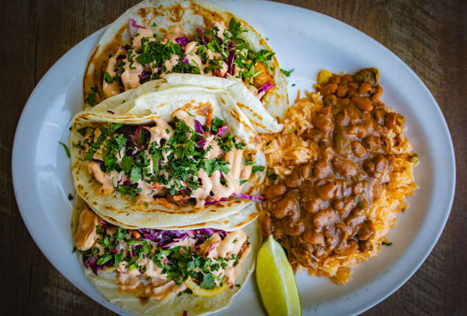 Top-down view of three Mexican-style soft tacos with creamy orange sauce, shredded purple cabbage and chopped cilantro, served with seasoned Mexican rice, pinto beans and a lime wedge on a white plate.