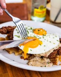 Close-up American brunch plate: chicken-fried steak and home fries topped with creamy gravy, two sunny-side-up eggs with oozing yolks and chives as a knife cuts the yolk