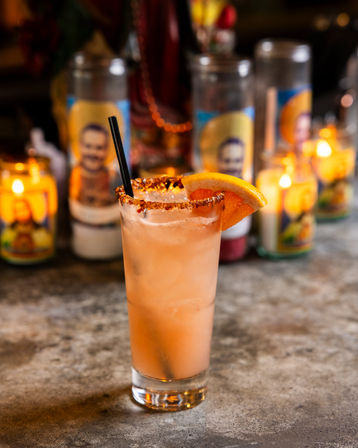 Pink grapefruit cocktail in a tall highball glass with chili-salt rim, grapefruit wedge and black straw on a concrete bar top, blurred glowing candles in the background.