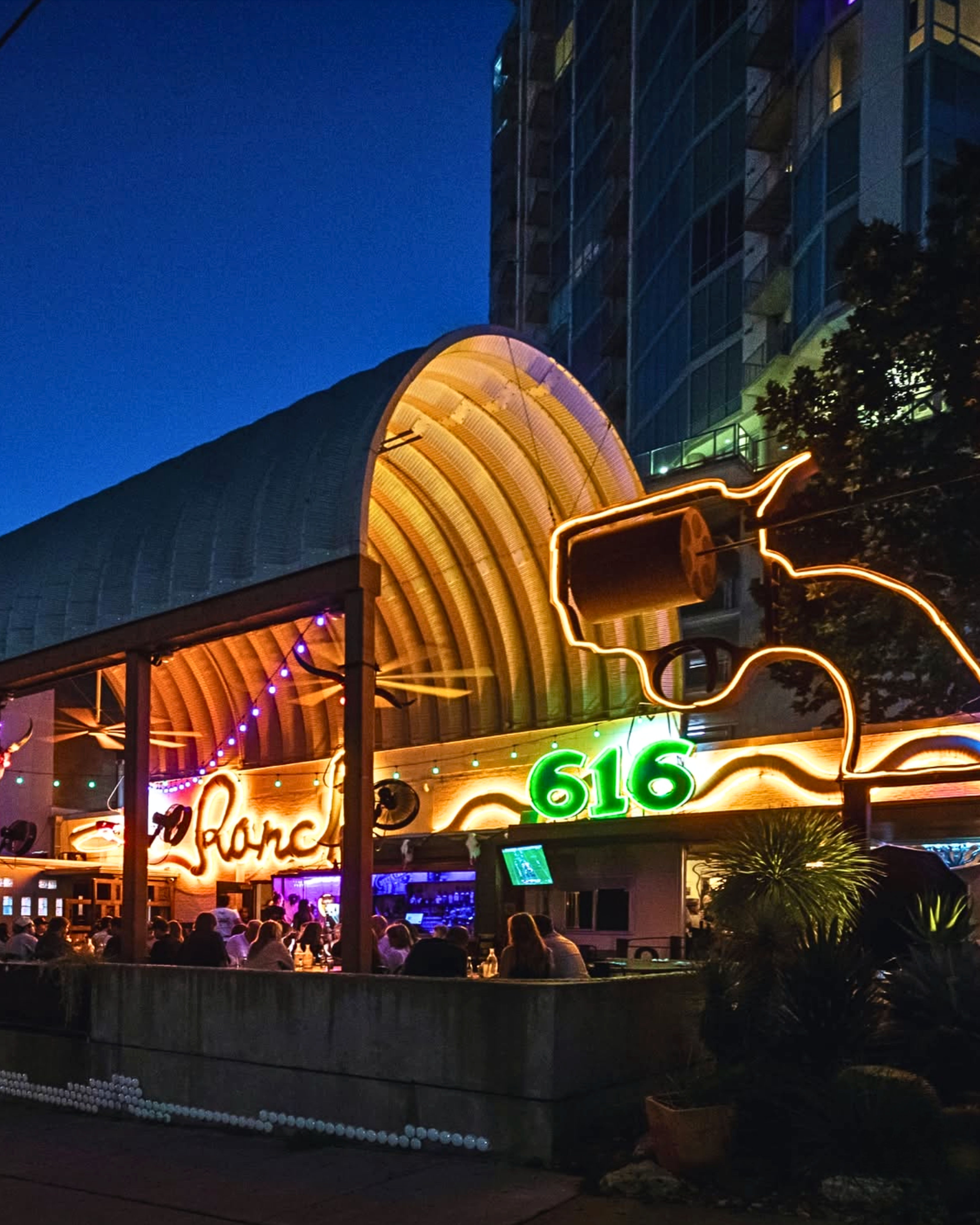 Outdoor nighttime bar under a curved wooden canopy with colorful neon '616' sign, string lights and a glowing outline sculpture, patrons dining in a lively urban setting.