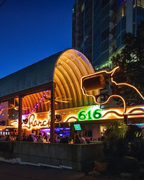 Outdoor nighttime bar under a curved wooden canopy with colorful neon '616' sign, string lights and a glowing outline sculpture, patrons dining in a lively urban setting.