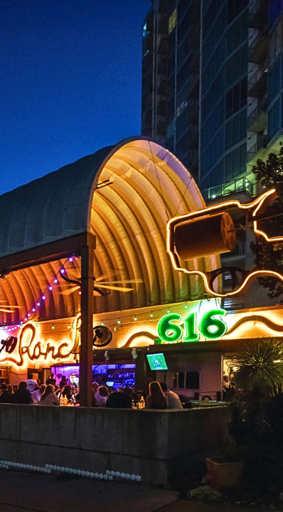 Outdoor nighttime bar under a curved wooden canopy with colorful neon '616' sign, string lights and a glowing outline sculpture, patrons dining in a lively urban setting.