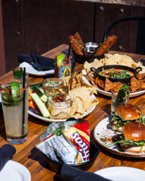 Sunny outdoor wooden table with shareable plates: tortilla chips and assorted dips in small jars, fried coated sticks with dipping sauces, mini brioche sliders, cucumber and veggie sticks, and spiced lime cocktails garnished with chili and lime.