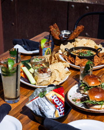 Sunny outdoor wooden table with shareable plates: tortilla chips and assorted dips in small jars, fried coated sticks with dipping sauces, mini brioche sliders, cucumber and veggie sticks, and spiced lime cocktails garnished with chili and lime.