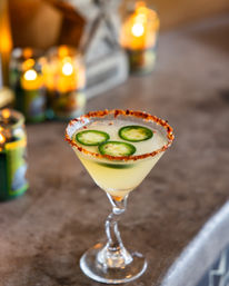 Chili-salt-rimmed jalapeño cocktail in a twisted-stem martini glass, topped with three jalapeño slices and set on a bar countertop with warm candles glowing in the background.